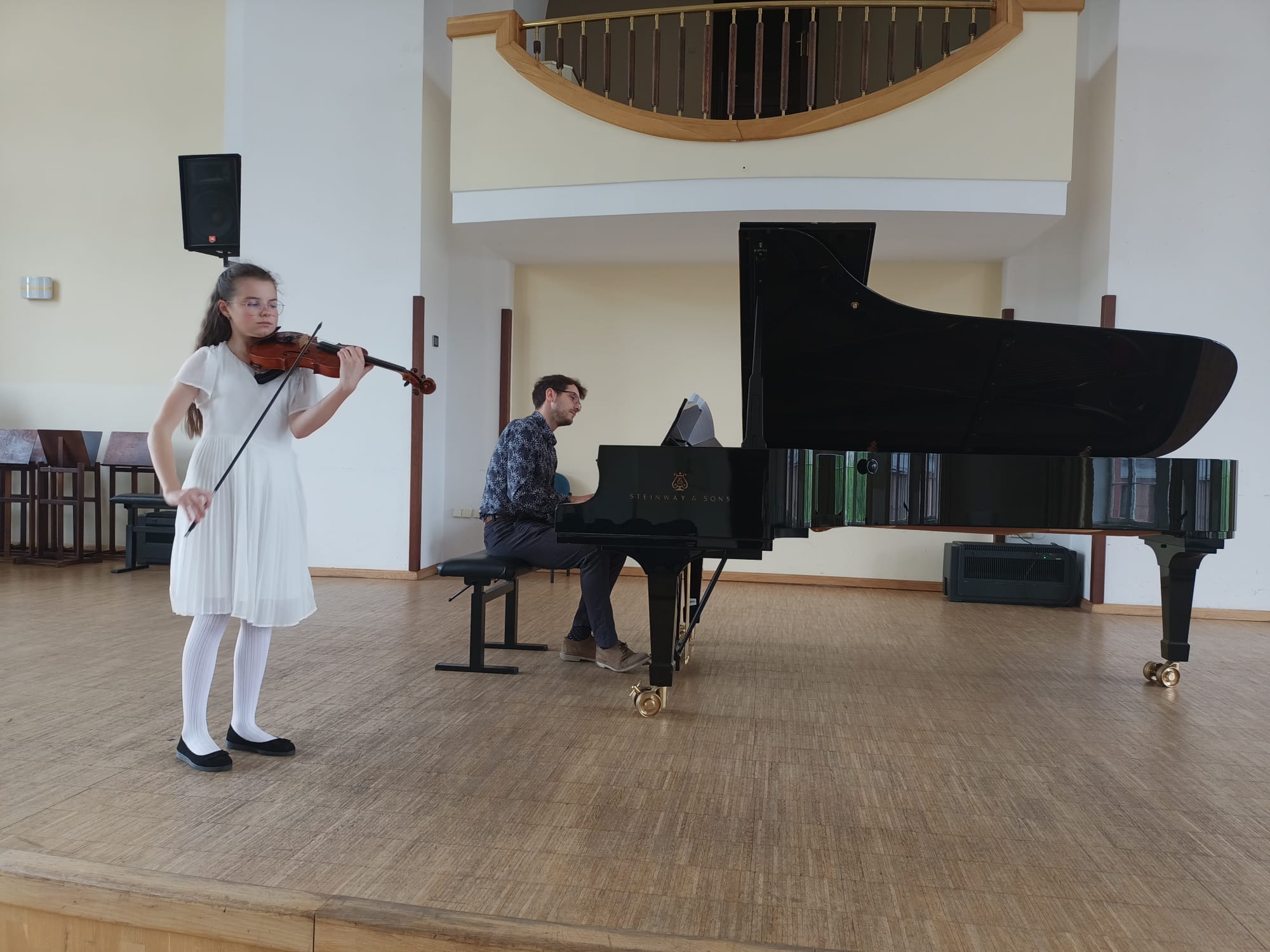 Na scenie po lewej stronie uczennica naszej szkoły w białej sukience grająca na skrzypcach. w Tle czarny fortepian,a przy nim akompaniator.