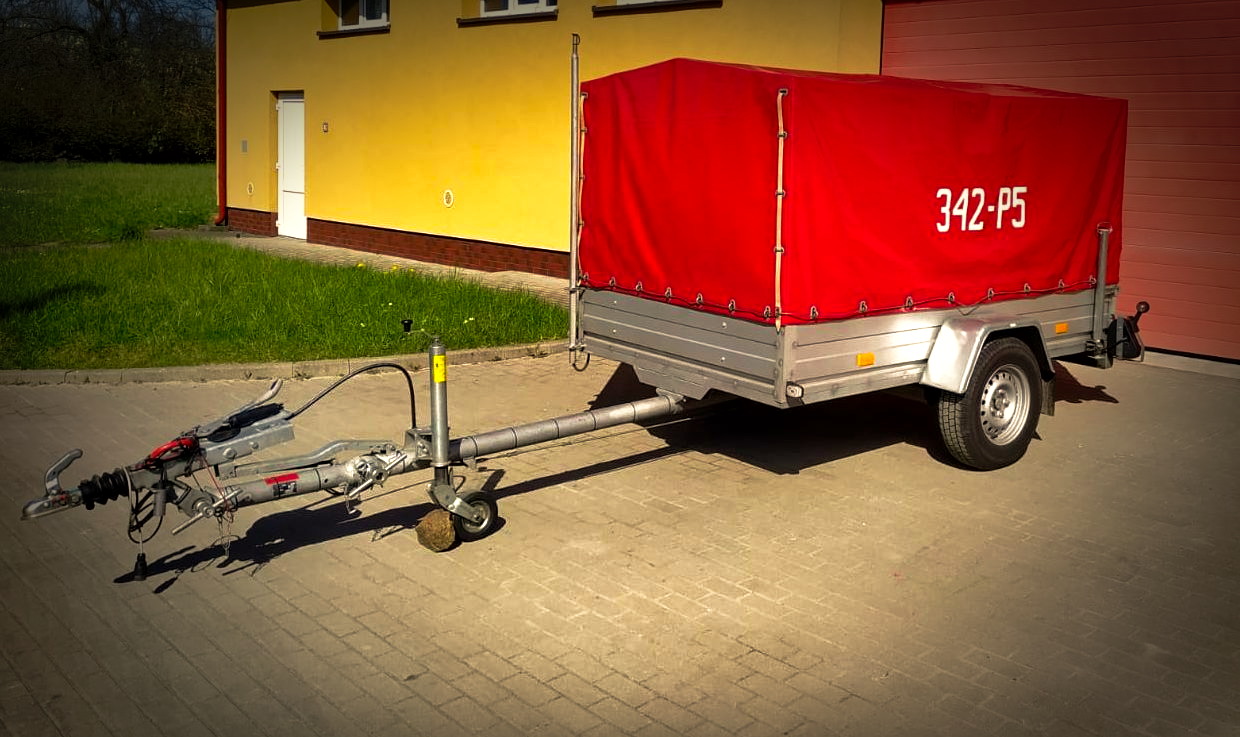 Zdjęcie przedstawia przyczepę wraz z plandeką koloru czerwonego z na tle bram garażowych przy jednostce ratowniczej nr 2 w Słupsk, 