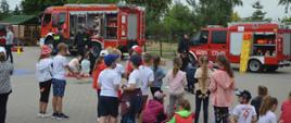 Boisko Zespołu Szkolno- Przedszkolnego nr 2 w Rawiczu. Na pierwszym planie dzieci przyglądające się pokazowi udzielania pierwszej pomocy w wykonaniu strażaków. W tle dwa samochody ratowniczo-gaśnicze.