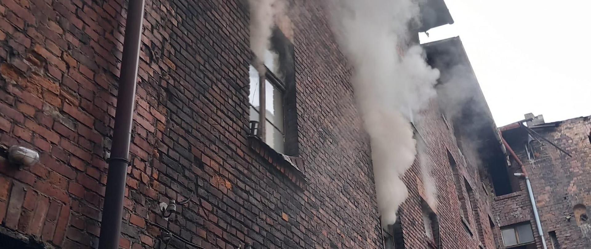 Na zdjęciu widoczny budynek, z okien na pierwszym piętrze wydobywa się dym