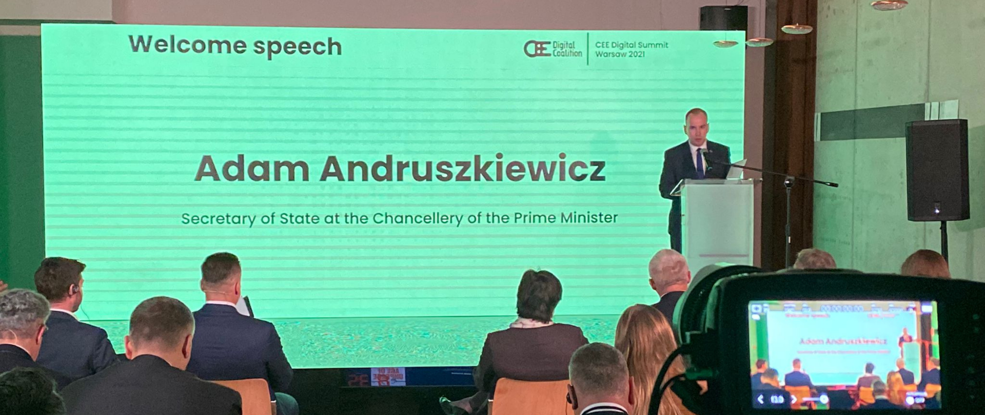 Minister Adam Andruszkiewicz podczas wystąpienia, za nim ekran z jego imieniem i nazwiskiem, przed nim - uczestnicy Szczytu.