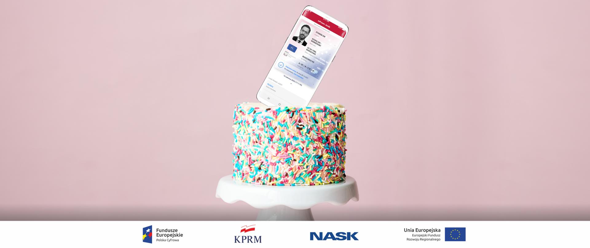 Kolorowy tort na jasnoróżowym tle, leży na białej paterze, na środku tortu - zamiast świeczki - telefon, na ekranie którego wyświetla się mPrawo Jazdy.