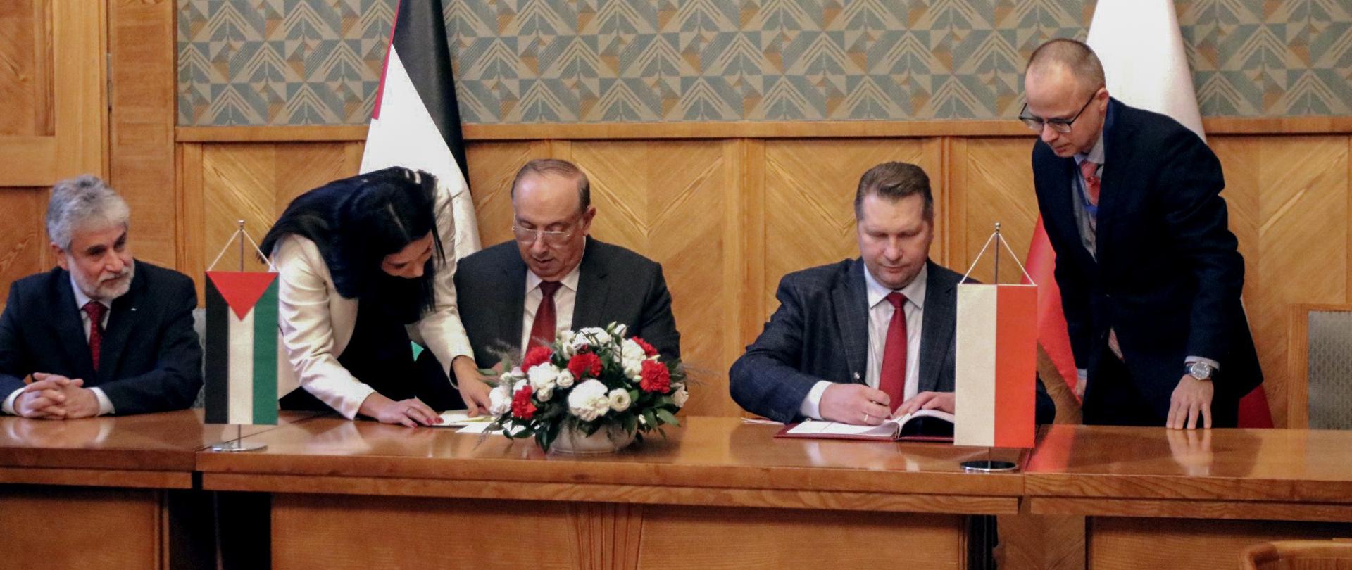 Za drewnianym stołem siedzi trzech mężczyzn w garniturach, jednemu z nich kobieta w białym ubraniu podaje dokumenty, przed nimi małe flagi Polski i Palestyny i bukiet biało-czerwonych kwiatów. Za nimi ściana w zielony wzorek.