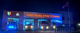 Wieczorem, w oświetlonych bramach garaży komendy Państwowej Straży Pożarnej stoją samochody ratowniczo - gaśnicze i specjalny drabina z włączonymi sygnałami świetlnymi, przed samochodami w szeregu stoją w ubraniach koszarowych funkcjonariusze oddając hołd strażakowi z Komendy Miejskiej PSP w Gdańsku. Po prawej stronie maszt z flagą państwową. Na budynku, nad grażami podświetlony napis Państwowa Straż Pożarna logo Państwowej Straży Pożarnej i herb miasta Żory.