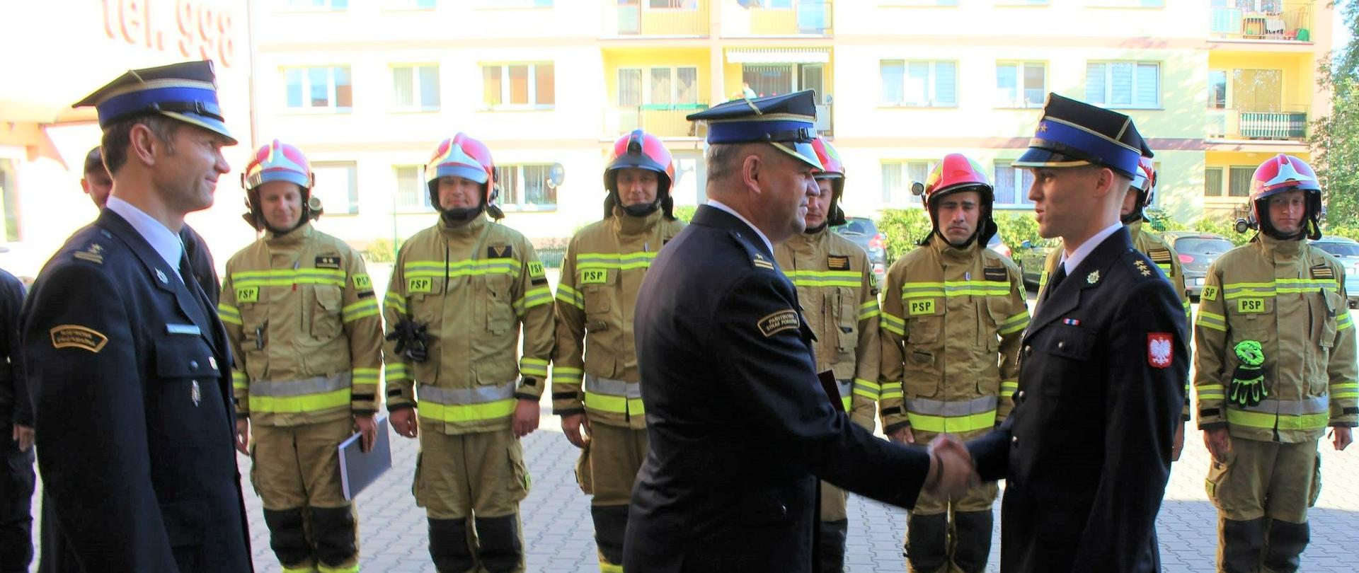Zdjęcie przedstawia uroczyste wręczenie decyzji nagrodowej dla strażaka.
W tle plac Komendy, inni strażacy i budynki.
