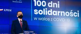 Premier Mateusz Morawiecki na tle ścianki wizyjnej z napisem: 100 dni solidarności w walce z COVID-19.