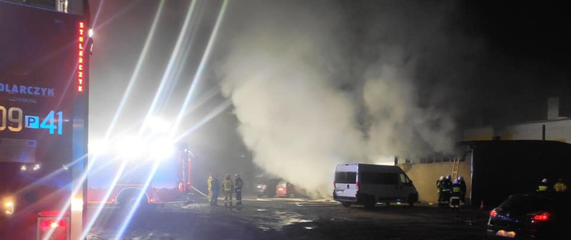 Zdjęcie przedstawia samochód strażacki oraz strażaków gaszących pożar w budynku myjni samochodowej.
W tle budynek i gęsty dym.
