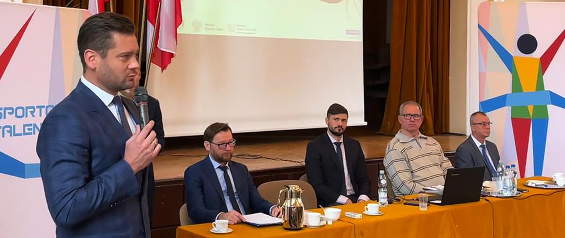 Minister Kamil Bortniczuk na spotkaniu z nauczycielami w Opolu - program Sportowe Talenty; minister stoi z mikrofonem, w tle stół przy którym siedzą prelegenci