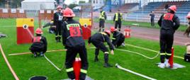 Na zdjęciu strażacy młodzieżowej drużyny pożarniczej podczas zawodów sportowo-pożarniczych. W tle sędziowie konkurencji oraz trybuny boiska.