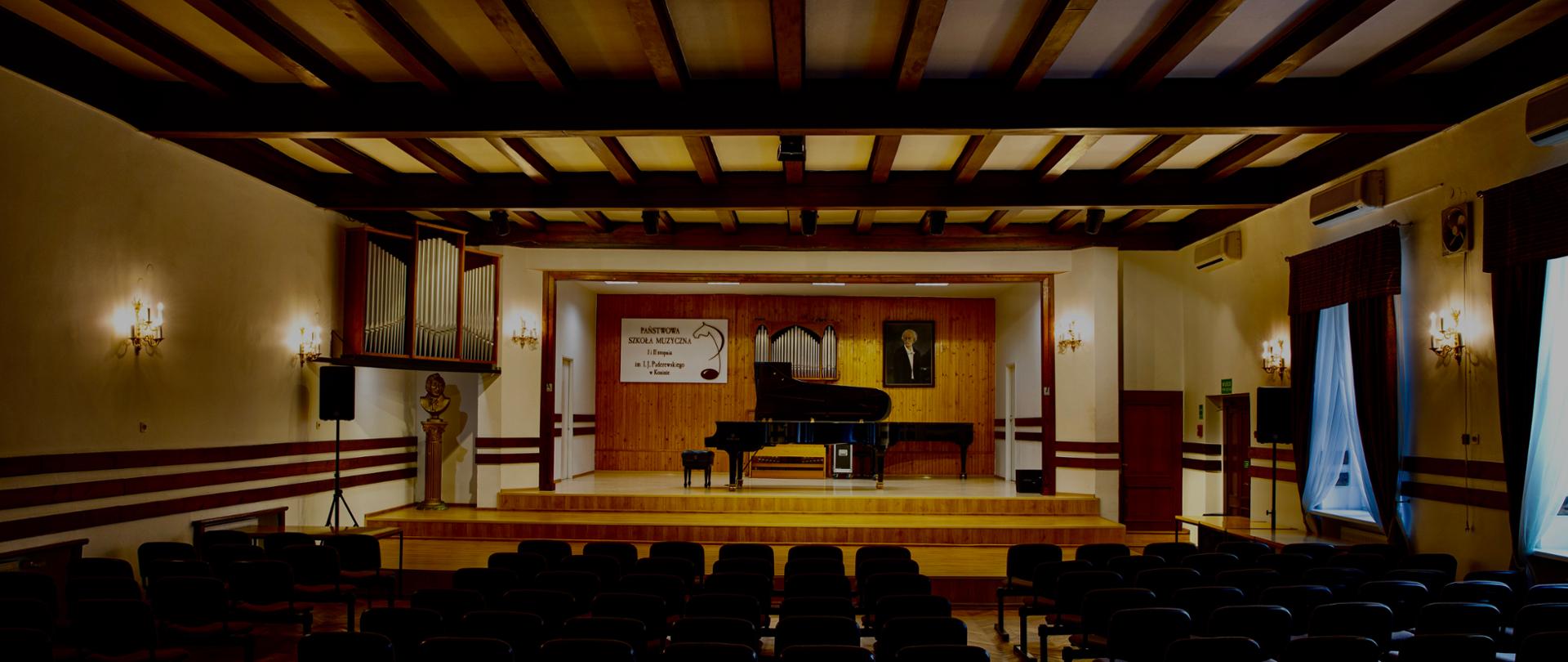 Sala koncertowa, widownia z krzesłami, na scenie dwa fortepiany, logo szkoły wisi z tyłu, portret I. J. Paderewskiego, ściany białe, scena kolor drewna.