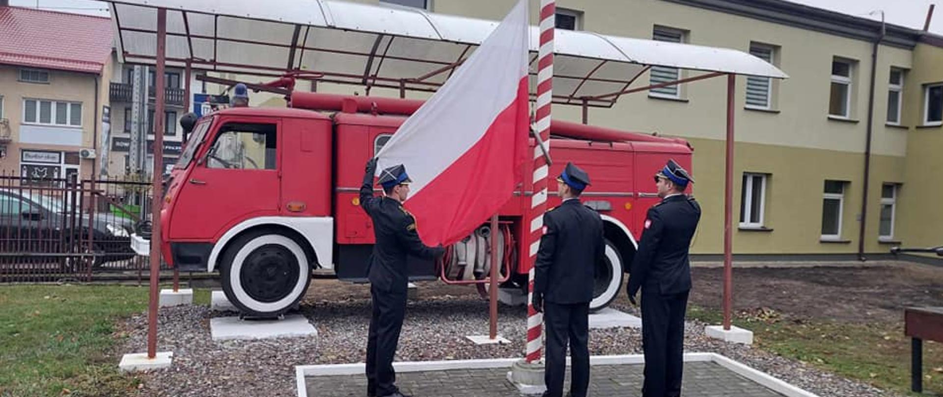 Poczet flagowy stoi przed masztem, do którego przyczepili flagę Polski. Jeden z funkcjoanriuszy prezentuje flagę. W tle widać zabytkowy samochód strażacki.
