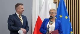 Minister Wieczorek i wiceminister Mrówczyńska stoją na sali, minister trzyma w ręku kartki papieru, wiceminister Mrówczyńska trzyma czerwoną teczkę i mówi do mikrofonu na stojaku, za nimi pod ścianą flagi Polski i UE.