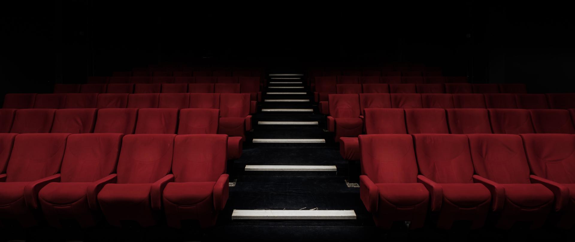 Zaciemniona sala kinowa z bordowymi fotelami i podświetlonymi schodkami po środku sali.