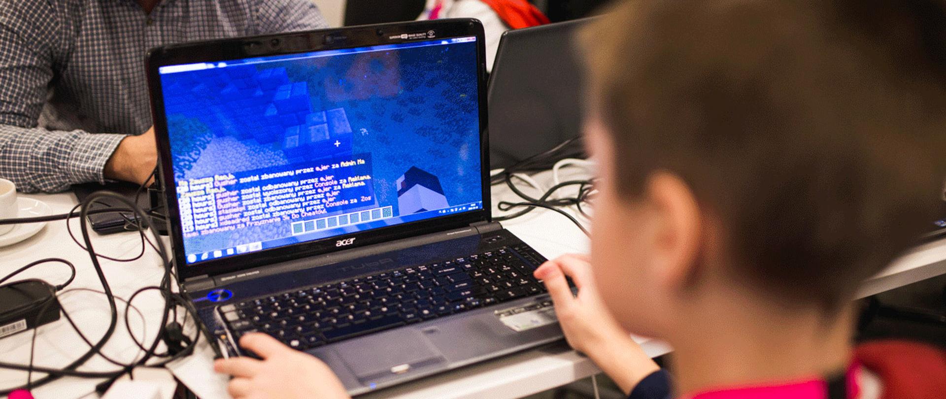 Na zdjęciu widać chłopca korzystającego z laptopa, którego ekran pokazuje popularną grę Minecraft. Przy tym samym stole siedzi przy laptopach więcej osób.
