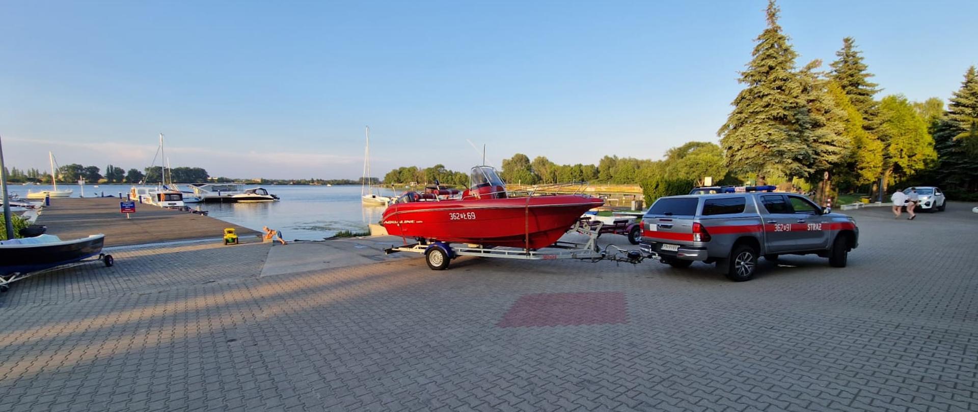 Zdjęcie przedstawia samochód strażacki z łodzią na przystani przed wodowaniem. Na zdjęciu widać pomost, łodzie oraz jezioro