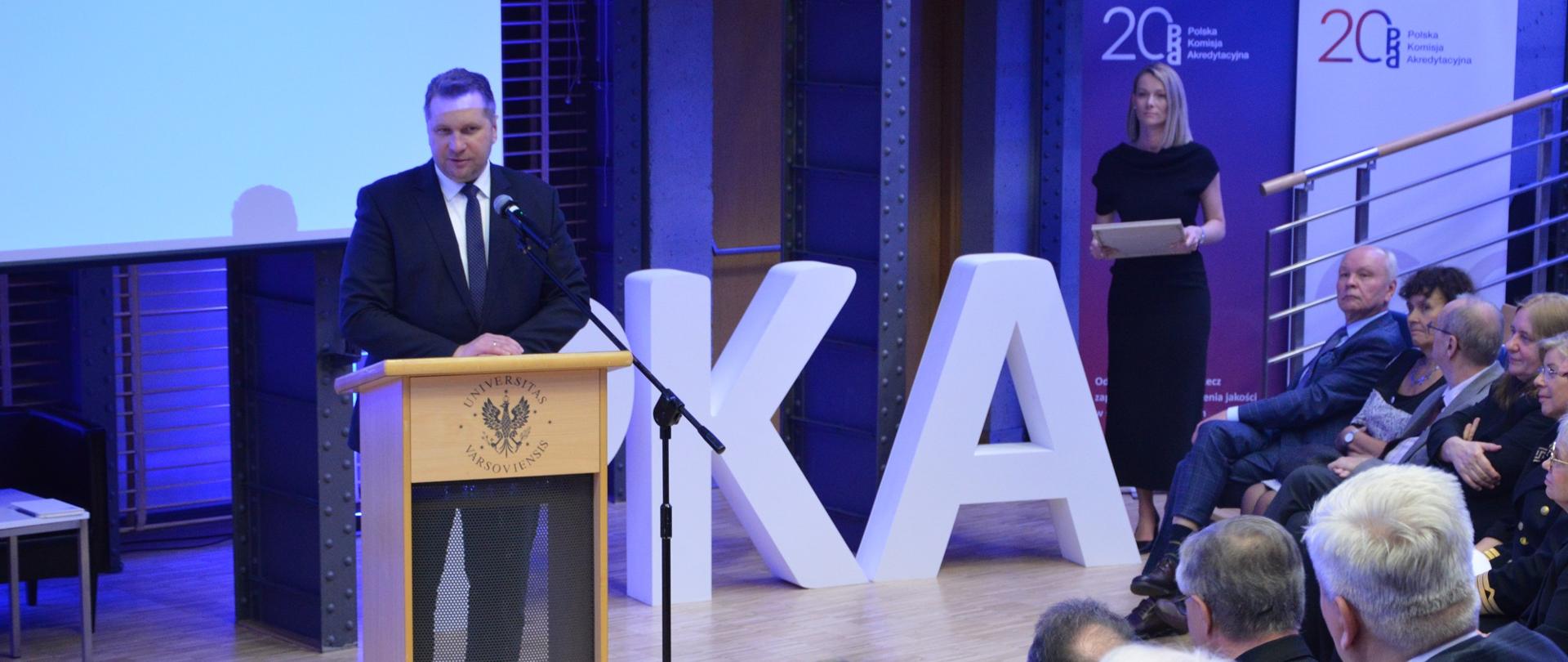 Minister Czarnek stoi przy mównicy i mówi do mikrofonu, przed nim na niebieskich krzesłach siedzą elegancko ubrani ludzie.