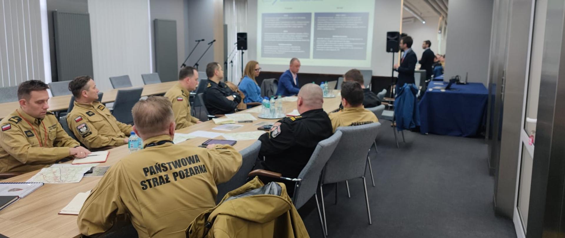 Uczestnicy spotkania siedzący przy stole. Ośmiu mężczyzn w mundurach Państwowej Straży Pożarnej. Jedna osoba stoi i prezentuje slajd na ekranie. 