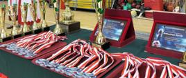 Na stole pokrytym płótnem leżą pamiątkowe medale, stoją puchary i dyplomy związane z mistrzostwami. Obok stołu stoją dwie kobiety w strojach ludowych