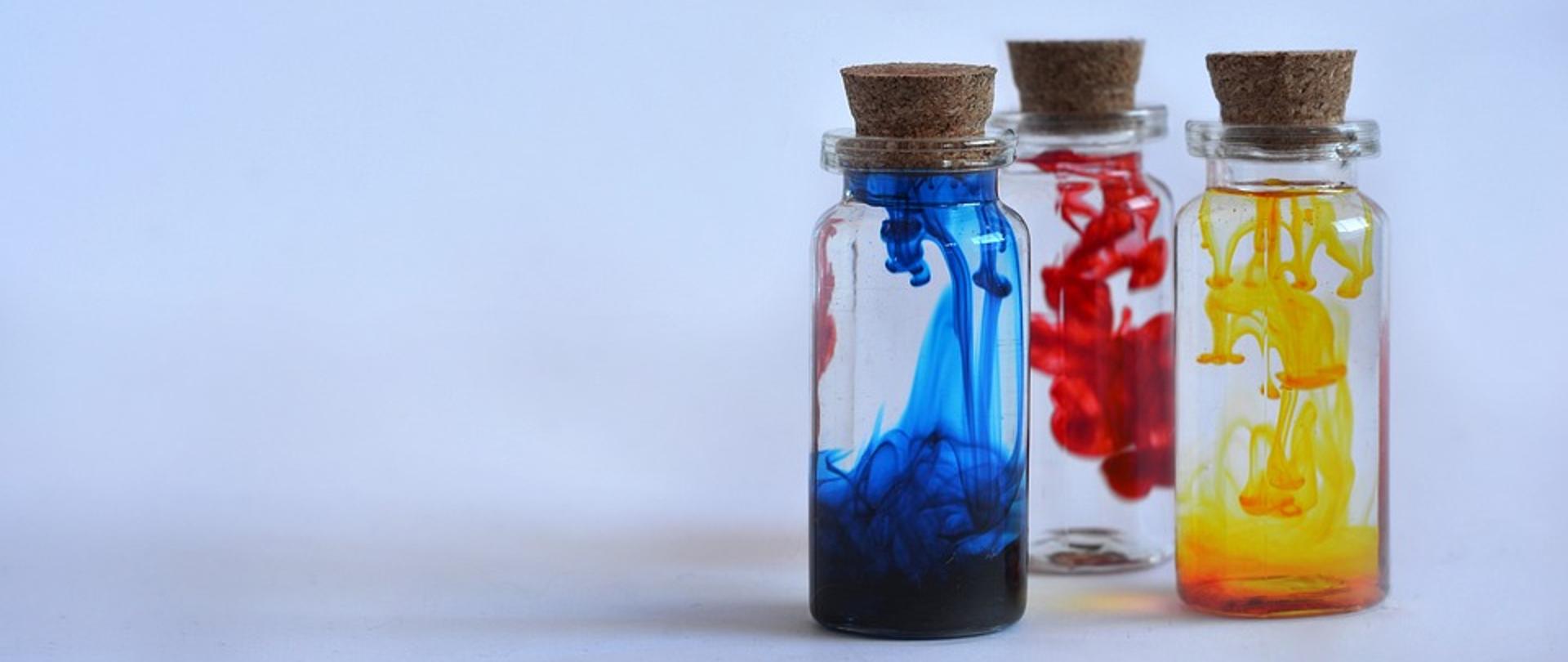 Trzy zakorkowane butelki z kolorową zawartością, z lewej strony z niebieskim płynem, w środku z czerwonym płynem, po prawej stronie z żółtym płynem