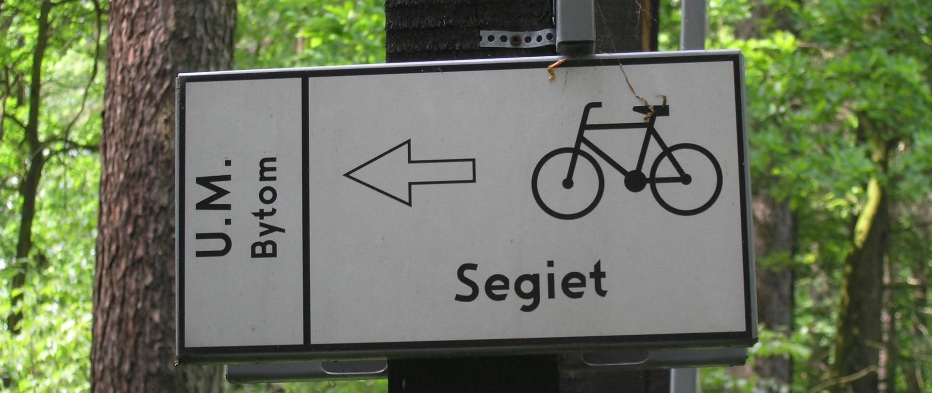 Zdjęcie przedstawia tabliczkę oznaczającym kierunek drogi rowerowej do rezerwatu Segiet, w tle las