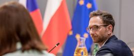 Wiceminister Waldemar Buda siedzi przy stole konferencyjnym na tle flag Polski, Czech i UE