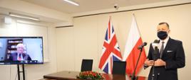 Na zdjęciu widać wysokiego mężczyznę, który jest ubrany w garnitur i stoi przy mikrofonie. W tle widać stół z kwiatami i flagi Wielkiej Brytanii i Polski. 