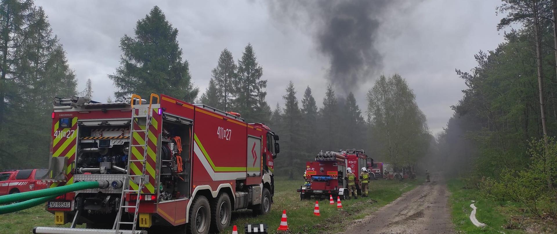 Na zdjęciu duży samochód strażacki przed nim kolejne. Dalej w oddali lasy nad którymi unosi się ciemny dym.