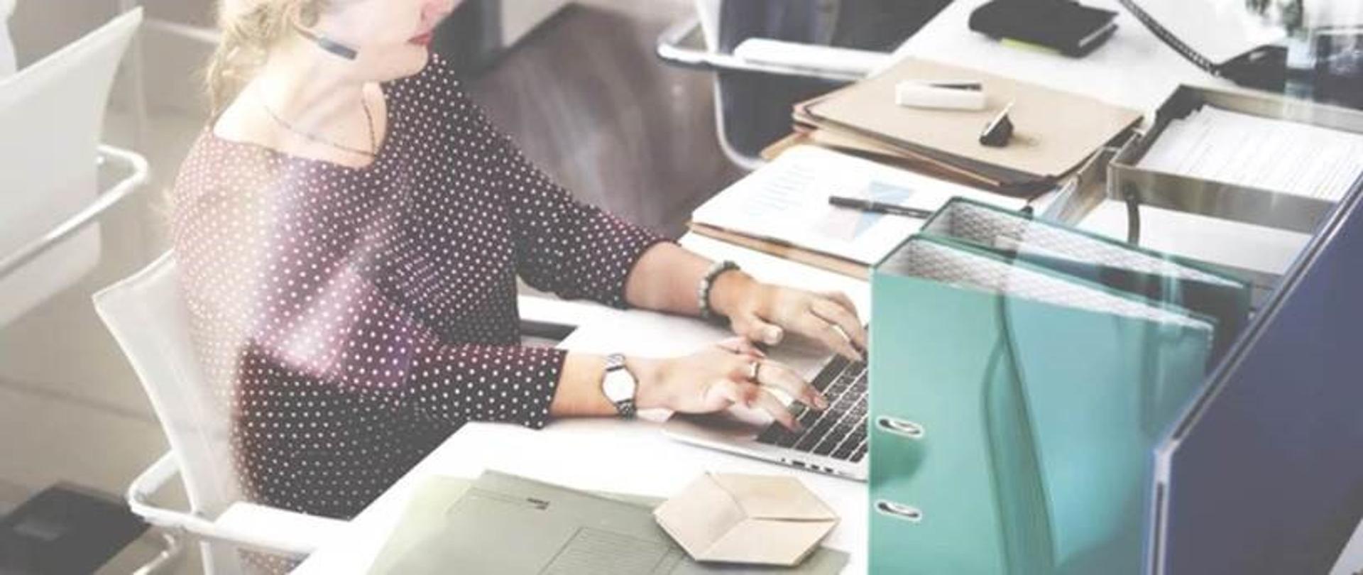 Kobieta w fioletowej bluzce w kropki, pisząca na laptopie przy biurku, rozmawiająca przez słuchawki 