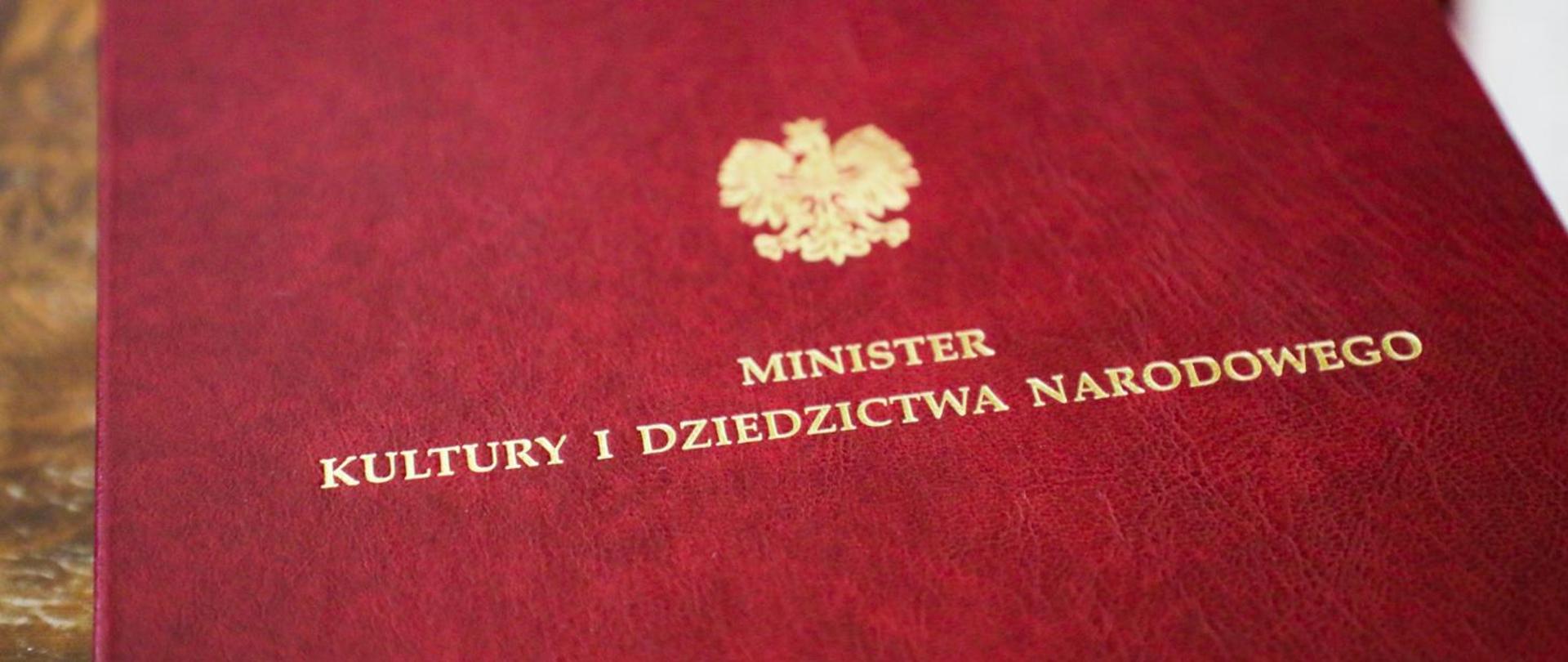 fragment czerwonej okładki ze złotym Godłem Polski oraz napisem Minister Kultury i Dziedzictwa Narodowego
