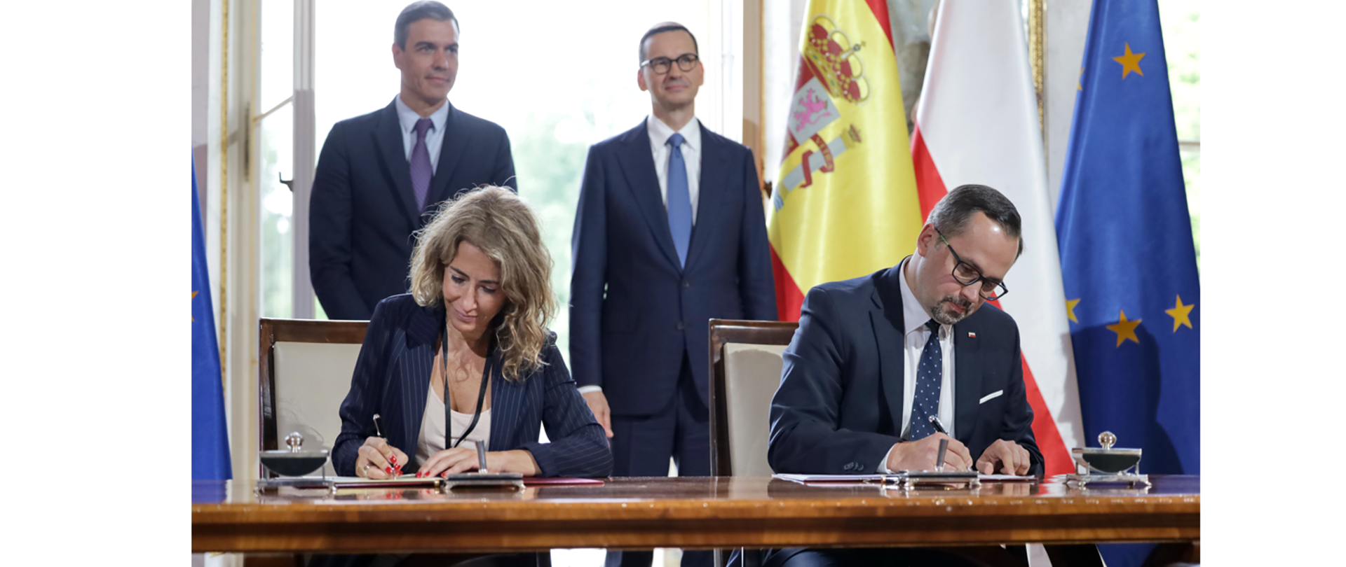 Marcin Horała i Raquel Sánchez Jiménez podpisuja przy stole dokumenty. Za nimi stoją premierzy Mateusza Morawieckiego i Pedro Sancheza.