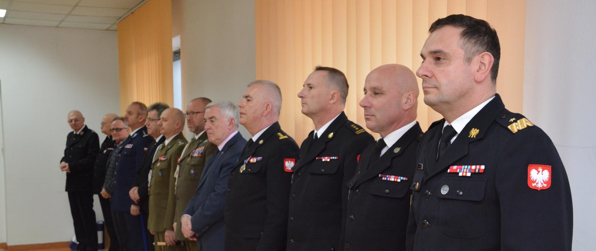 Na zdjęciu widzimy mężczyzn - przedstawicieli różnych służb mundurowych w umundurowaniu oraz przedstawicieli władz administracyjnych w garniturach.