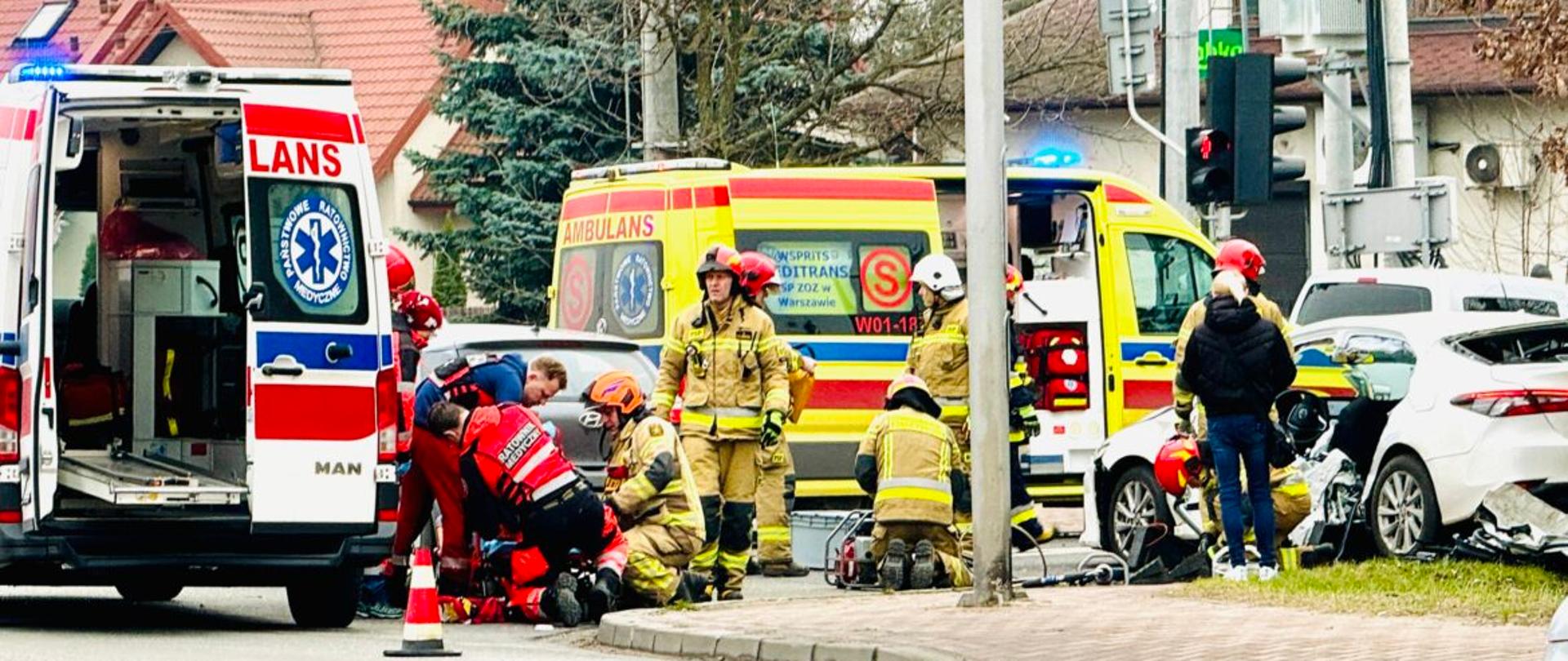 Na zdjęciu widać strażaków i ratowników medycznych udzielających pomocy poszkodowanym w wypadku ma tle karatek pogotowia.