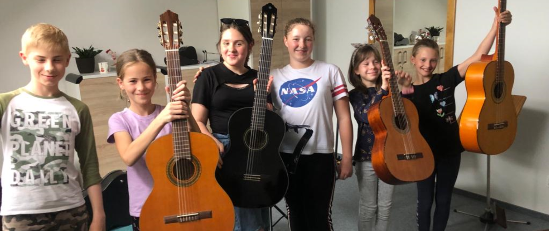 Na zdjęcie sześcioro uczniów (pięć dziewczyn i jeden chłopak), Wszyscy są uśmiechnięci i trzymają gitarę. W tle za nimi komoda i szafa w kolorystyce: front w kolorze brązu, korpusy białe, po boku wisi lustro.