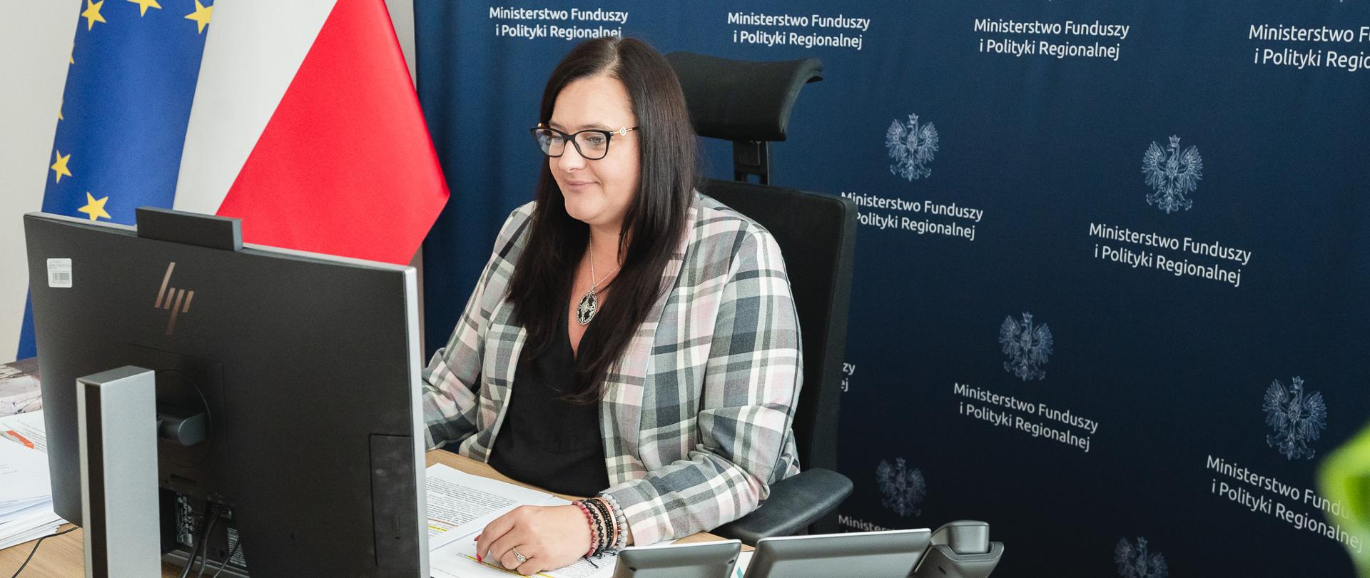 Wiceminister Małgorzata Jarosińska-Jedynak siedzi na fotelu przy biurku i patrzy na monitor, który stoi przed nią. W tle z lewej strony flagi Unii Europejskiej i Polski.