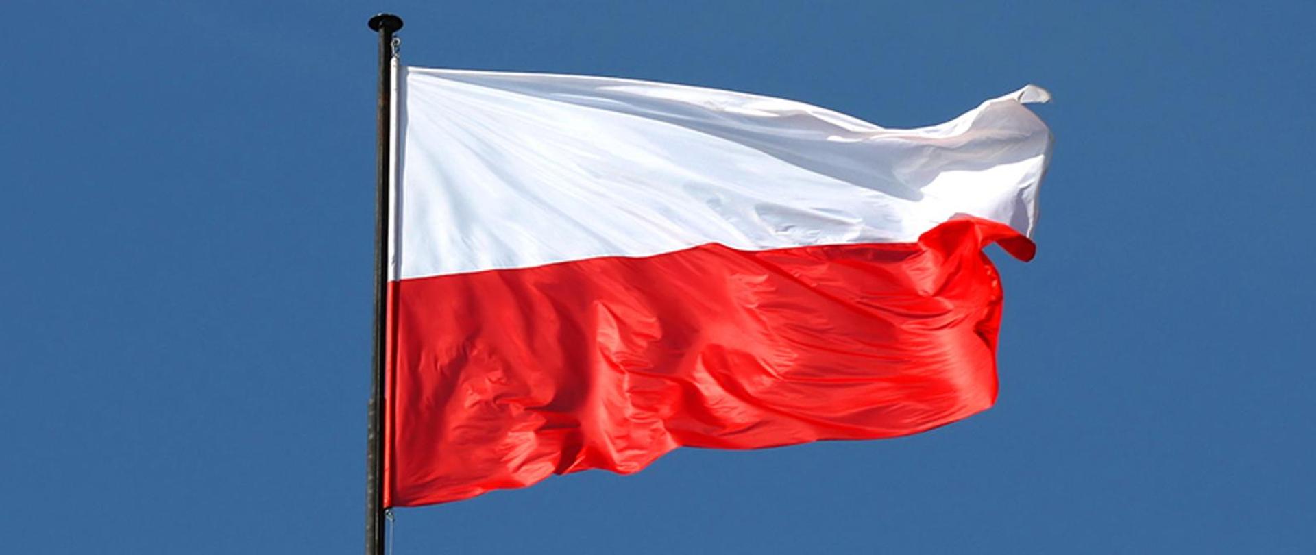 Zdjęcie przedstawia biało-czerwoną Flagę RP na maszcie. Flaga na niebieskim tle.