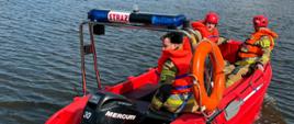 Na łódce ratowniczej płynie 3 strażaków ubranych w stroje strażackie, kamizelki ratunkowe. 