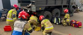 Widoczni strażacy ćwiczący wydobycie manekina ćwiczebnego znajdującemu się pod kołami ciężarówki.