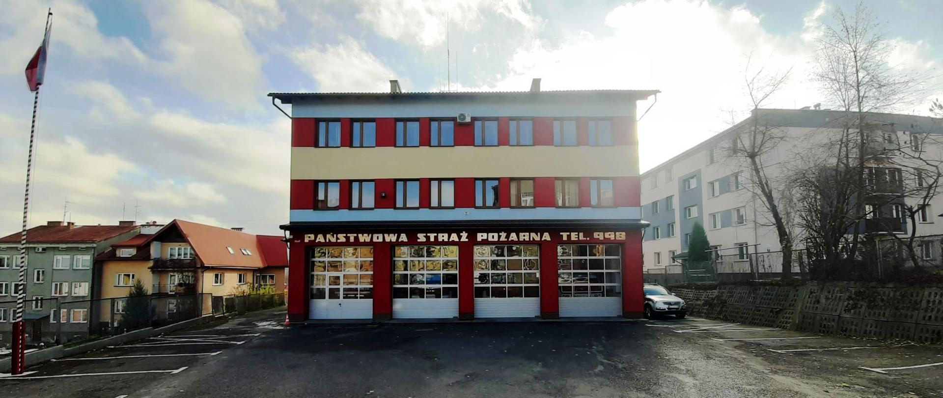 Fotografia kolorowa przedstawia widok trzykondygnacyjnego budynku Komendy Powiatowej Państwowej Straży Pożarnej w Brzozowie. Na pierwszym planie asfaltowy plac manewrowy przed budynkiem. Po lewej stronie na maszcie flagowym powiewa biało-czerwona flaga. Na środku drugi plan wypełnia ściana frontowa budynku Komendy Powiatowej Państwowej Straży Pożarnej w Brzozowie, malowana pasami koloru czerwonego, niebieskiego i żółtego, w okresie jesiennym. Na ścianie parteru napis o treści Państwowa Straż Pożarna Tel. 998. Na parterze widoczne cztery srebrne bramy garażowe, na pozostałych dwóch piętrach dwa rzędy okien.