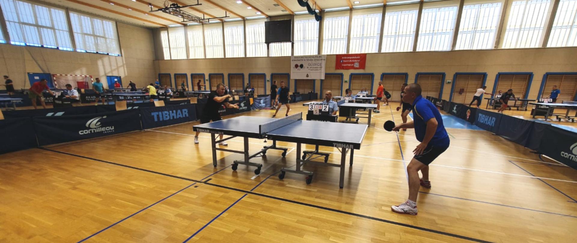 Na zdjęciu widoczne wnętrze hali sportowej, na pierwszym stół do tenisa, przy których grają zawodnicy w strojach sportowych. Na dalszym planie kolejne stoły i grający zawodnicy.