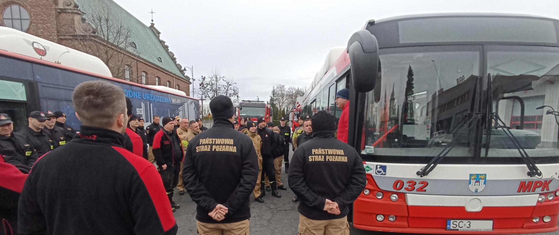 Na zdjęciu dwa autobusy miejskie obok nich ustawieni strażacy w umundurowaniu służbowym