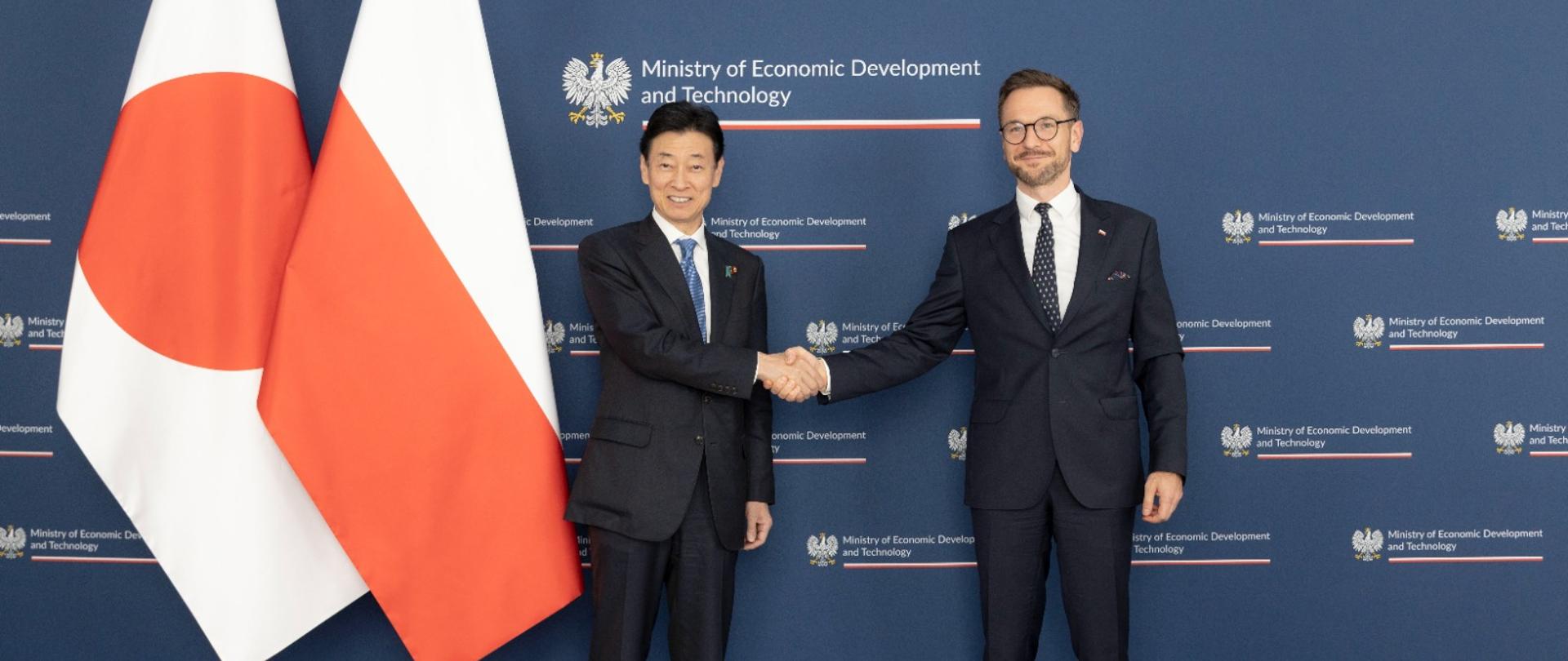 Spotkanie ministra Waldemara Budy z ministrem gospodarki, handlu i przemysłu Japonii, Yasutoshi Nishimura. Na zdjęciu ministrowie na ściance. Podają sobie rękę. Po lewej stronie flaga Japonii i Polski. 