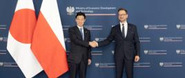 Spotkanie ministra Waldemara Budy z ministrem gospodarki, handlu i przemysłu Japonii, Yasutoshi Nishimura. Na zdjęciu ministrowie na ściance. Podają sobie rękę. Po lewej stronie flaga Japonii i Polski. 