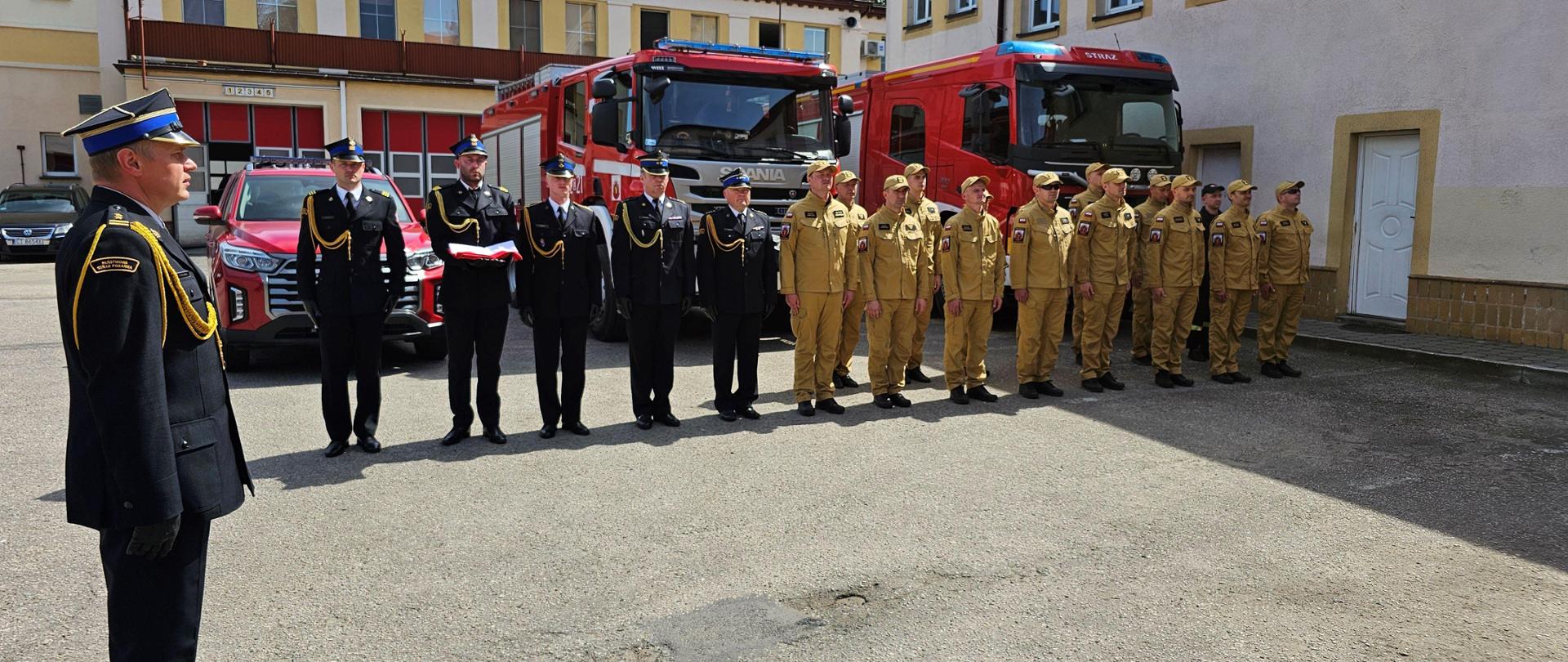 Zbiórka strażaków w piaskowych i czarnych mundurach na tle czerwonych pojazdów pożarniczych.
