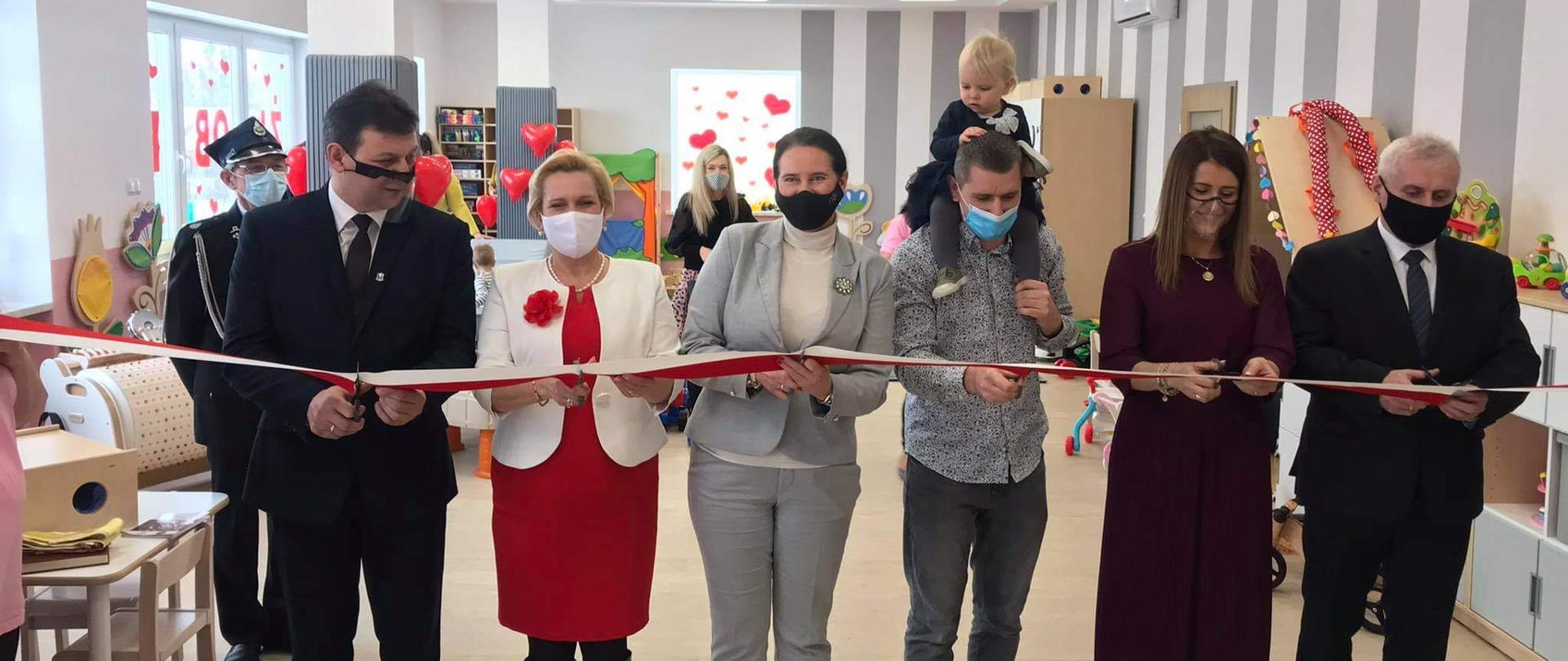 Uroczyste otwarcie żłobka w Gorzowie Śląskim. Na zdjęciu sześć osób, przecinające biało-czerwoną wstęgę na znak otwarcia obiektu. W tle sala żłobka. 