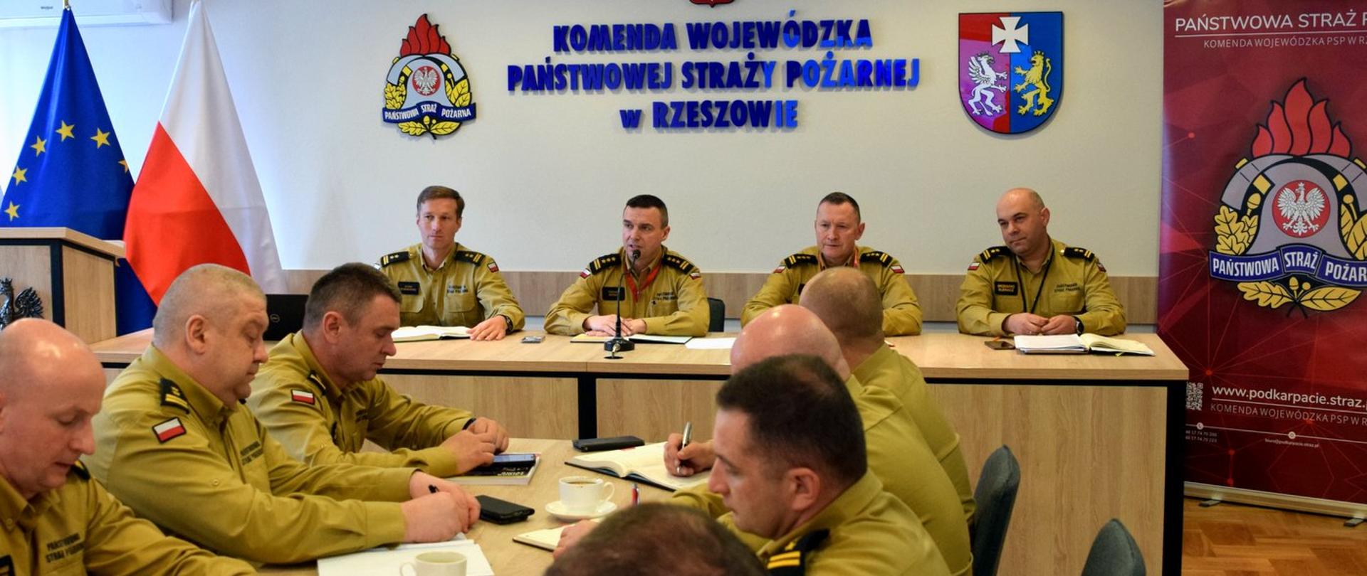 Zdjęcie zrobione w pomieszczeniu, podczas narady służbowej kadry kierowniczej Państwowej Straży Pożarnej. Na zdjęciu oficerowie PSP w piaskowych mundurach służbowych siedzą przy stole konferencyjnym.
