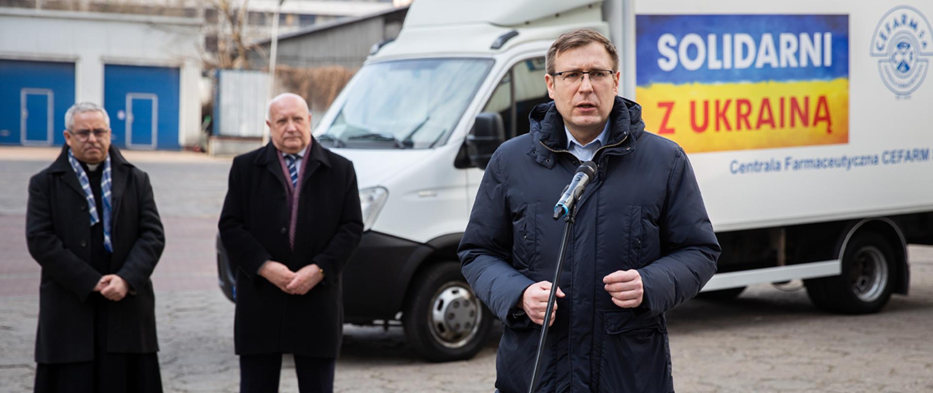 Wiceminister Maciej Małecki stoi przed mikrofonem. W tle biała ciężarówka z darami dla Ukrainy. Na ciężarówce żółto niebieska flaga z napisem "Solidarni z Ukrainą". Dodatkowo w tle stoi dwóch innych mężczyzn.
