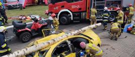 Strażacy podczas ćwiczenia ratowniczego. Z przodu zdjęcia żółty samochód osobowy przygnieciony drzewem, w tle samochód pożarniczy, quad strażacki, czerwone namioty wystawowe z napisami STRAŻ. 