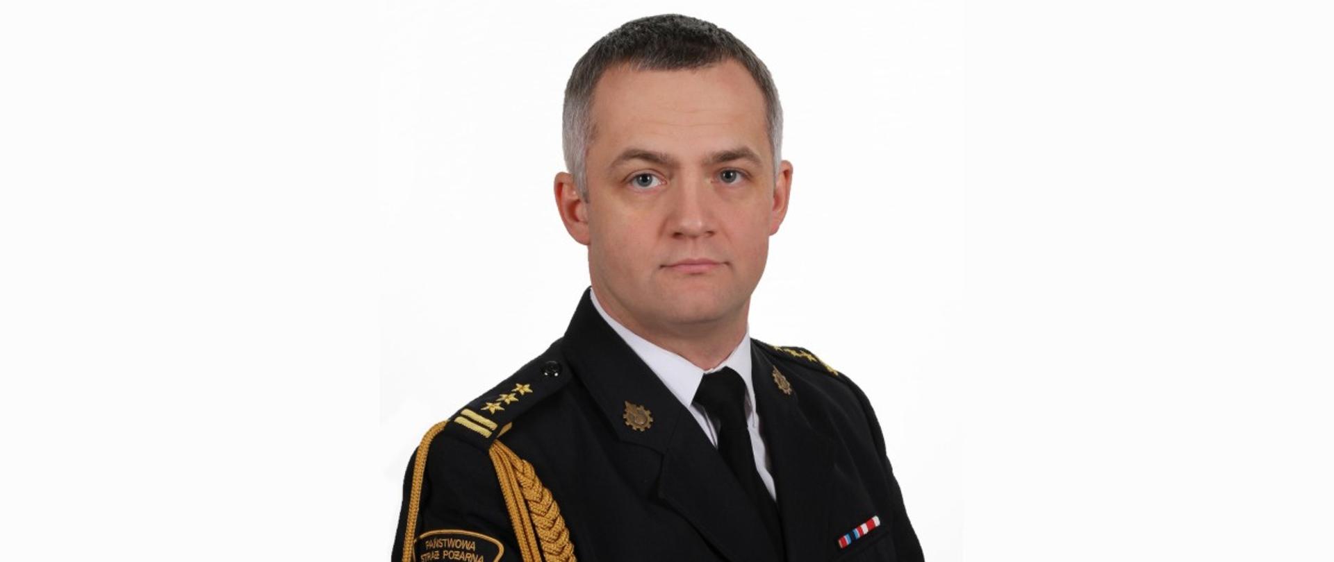 Zdjęcie przedstawia zdjęcie pełniącego obowiązki Zastępcy Komendanta Miejskiego Państwowej Straży Pożarnej bryg. Łukasza Szymańskiego ubranego w mundur galowy. Zdjęcie wykonane na białym tle.