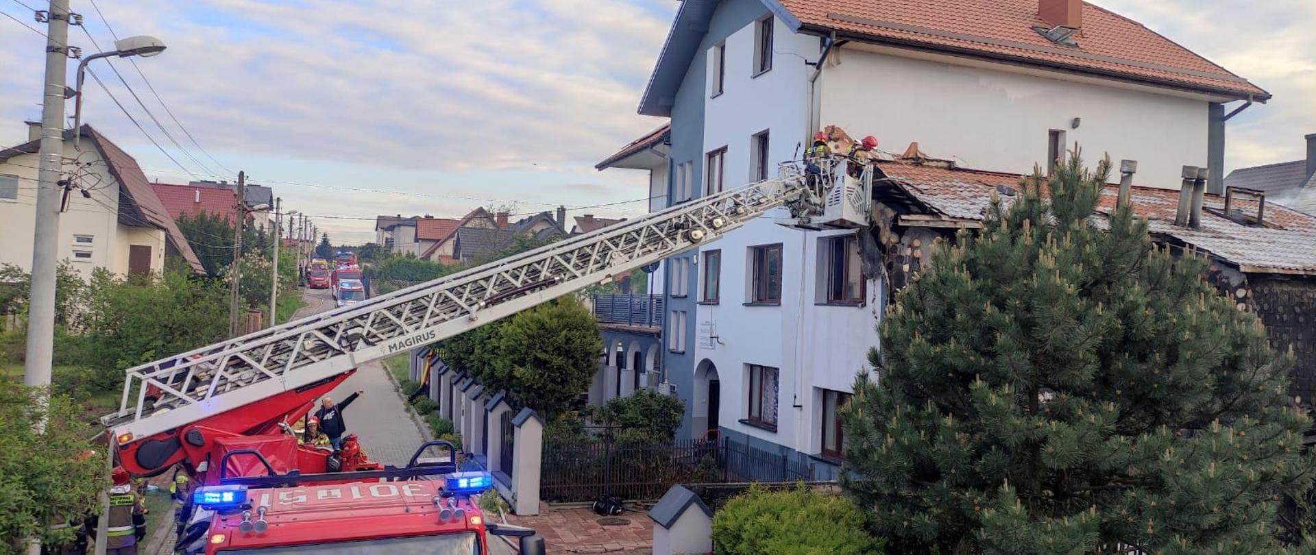 Zdjęcie przedstawia po prawej stronie budynek pensjonatu gdzie wybuchł pożar. Do budynku przystawiona drabina mechaniczna a w jej koszu ratownicy demontują elementy budynku.