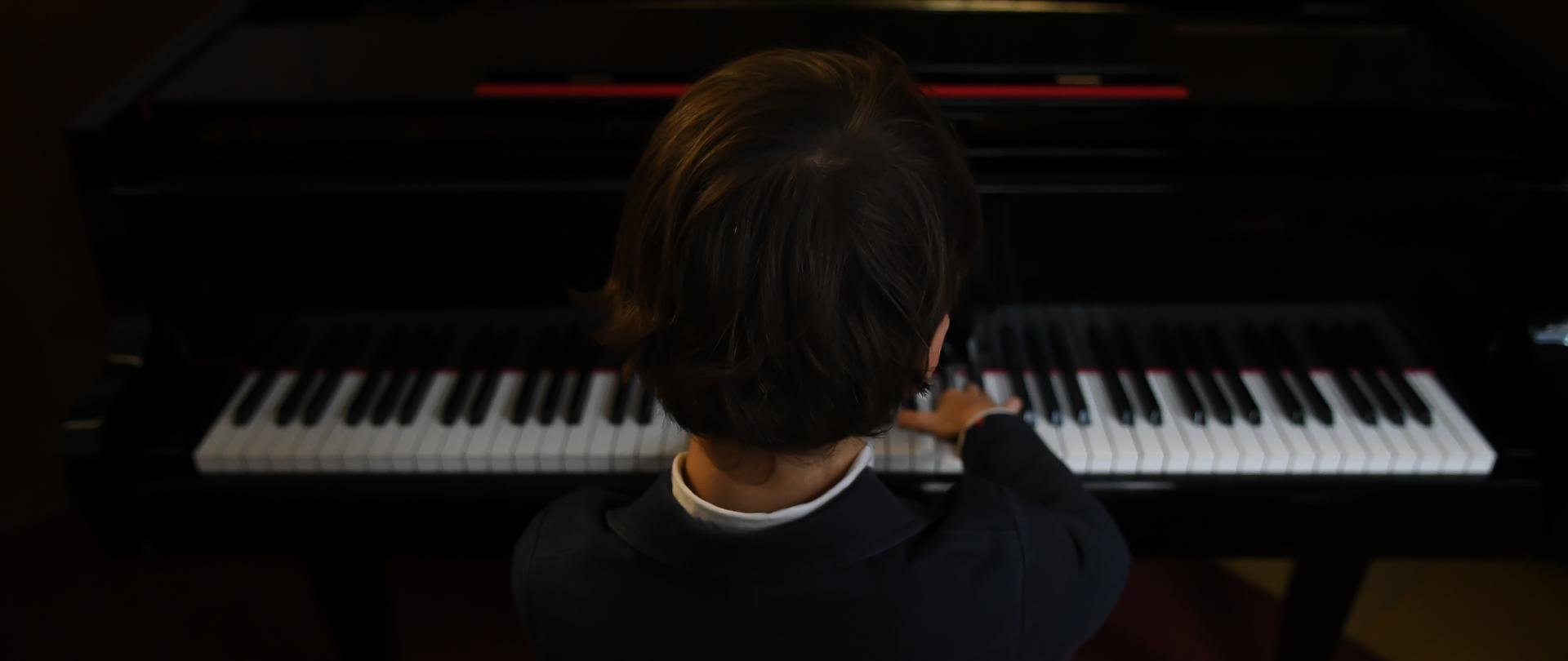 fotografia przedstawia ciemnowłosego chłopca siedzącego przy fortepianie ubranego w ciemny sweter 